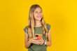 Excited blonde kid girl wearing green T-shirt over yellow studio background winking and eye hold smart phone use read social network news