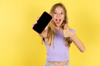 © Roquillo - Portrait blonde kid girl wearing violet T-shirt over yellow studio background holding in hands cell showing giving black screen thumb up