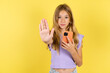 © Roquillo - blonde kid girl wearing violet T-shirt over yellow studio background using and texting with smartphone with open hand doing stop sign with serious and confident expression, defense gesture