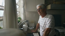 Thoughtful Senior Man Gazing Out Of Kitchen Window, Leaning On Counter. Retired Older Person Thinking While Staring Out Window