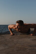 © gstockstudio - Strong young shirtless man doing push-ups while exercising outdoors with the sea on background