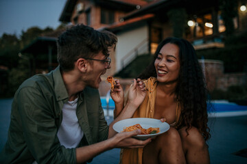 Wall Mural - Loving diverse couple sitting outdoors in the house yard during an evening and sharing a plate of pasta spaghetti.