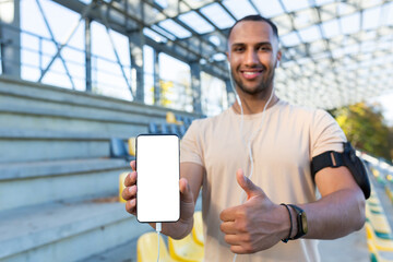 Young hispanic sportsman smiling and looking at camera, man in stadium showing white phone screen, while jogging and doing active exercise