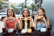 Three joyful smiling teenage girls (14-15 years old) sitting at a cafe outdoors, enjoying hamburgers and cola (coke) fresh drinks. Young ladies holding burgers, ready to eat them with pleasure.