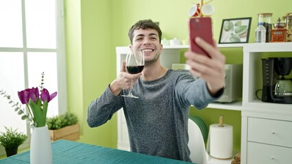 Wall Mural - Young hispanic man drinking glass of wine taking a selfie with smartphone at dinning room