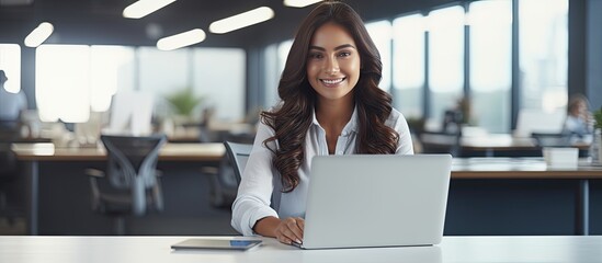 Female entrepreneur working at desk in office smiling at camera with empty laptop screen space for copy