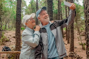 Wall Mural - Cheerful senior couple in a trekking day in the woods looking at smartphone to get a selfie, retired seniors man and woman and healthy lifestyle in nature