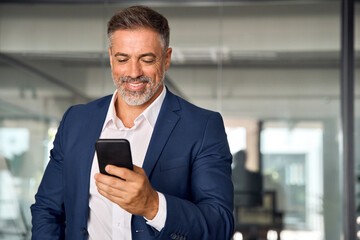 Wall Mural - Smiling mature Latin or Indian businessman holding smartphone in office. Middle aged manager using cell phone mobile app. Digital technology application and solutions for business success development.
