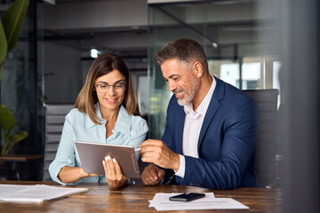 Team of diverse partners sitting at table mature Latin business man and European business woman discussing project on tablet in office. Two colleagues of professional business people working together.