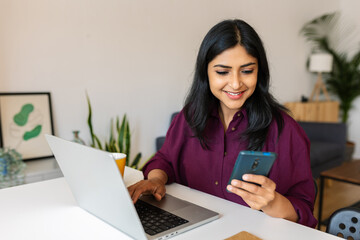 Happy young indian woman sitting on living room table at home working on laptop computer and using cell phone. Multitasking and business concept