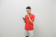 a young man wearing a red shirt on a white background with a cheerful expression while carrying a smartphone