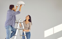 Happy Loving Couple Changing Light Bulb In New Apartment. Family Moving In New House, Doing DIY Renovations At Home. Cheerful Husband And Wife Standing On Ladder
