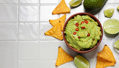 Wall Mural - Bowl of delicious guacamole with chili pepper, nachos chips and lime on white tiled table, flat lay