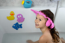 A Little Smiling Girl Of European Appearance With A Funny Bathing Hat Sits In The Bathroom With Toys. Care Of Children. Bathing A Child. Children's Hygiene