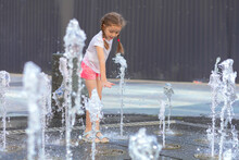 Active Little Girl Playing With Splash Spray Water. Happy Kid In Fountain In Wet Clothes. Summer Children Pastime, Entertainment, Recreation. Child's Leisure, Childhood In City