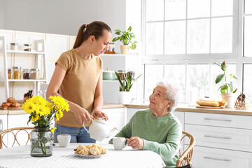 Wall Mural - Young woman pouring tea into cup for her grandmother in kitchen