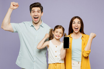 Wall Mural - Young parents mom dad with child kid girl 6 years old wearing blue yellow casual clothes use blank screen mobile cell phone do winner gesture isolated on plain purple background. Family day concept.