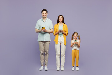 Wall Mural - Full body young happy parents mom dad with child kid daughter girl 6 year old wear blue yellow casual clothes hold in hand use mobile cell phone isolated on plain purple background Family day concept