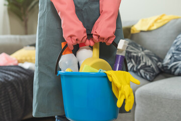 Wall Mural - Tired household clean up, housekeeper asian young woman, girl in apron, hand holding bucket of cleaning equipment, cleaning in living room at home. Messy maid or housewife organizing dirty and untidy.