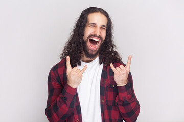 Carefree joyful man in checkered red shirt makes rock n roll gesture, heavy metal sign, enjoys favorite music, spends free time on party. Indoor studio shot isolated on gray background.