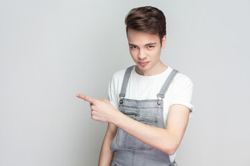 Get out. Portrait of serious angry young brunette man standing pointing finger, sneak male blaming another people, wearing denim overalls. Indoor studio shot isolated on gray background.