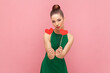 Portrait of winsome adorable woman with bun hairstyle standing showing little red hearts on stick, sending air kiss, wearing green dress. Indoor studio shot isolated on pink background.