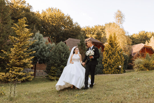 A stylish groom in a black suit and a cute bride in a white dress with a long veil are hugging and walking near green tall trees. Wedding portrait of smiling and happy newlyweds.