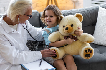 Kind senior female paediatrician doctor examining little child, cute girl during home visit or clinic check up. Concept of healthcare, medical assistance, insurance. Prescribing treatment for disease