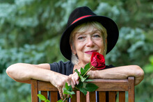 A Happy Aged Woman, Wearing A Black Hat, Holding A Gifted Red Rose Against The Background Of Nature.
