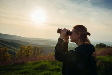 A hiker girl looks through binoculars at nature and birds standing on the top of a mountain in the bright rays of setting sun against the background of beautiful spring nature and cloudy blue sky