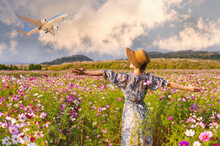 Beautiful Autumn Background. A Woman In A Floral Dress And Hat Stands In A Field Of Cosmos, Arms Wide Open, Watching An Airplane Take Off. 
