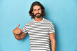 © Asier - Bearded man in a striped shirt, blue backdrop person pointing by hand to a shirt copy space, proud and confident