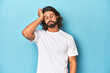 Bearded man in a white shirt, blue backdrop tired and very sleepy keeping hand on head.