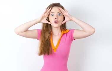 Wall Mural - Young woman wearing pink dress with scared expression, keeps hands on head, jaw dropped, has terrific expression. Omg concept