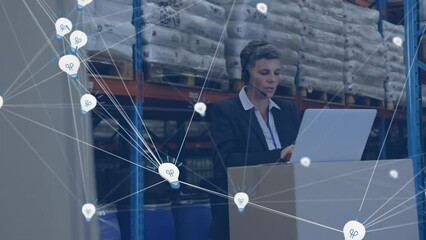 Canvas Print - Animation of network of bulb icons over caucasian female supervisor using laptop at warehouse