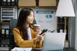 © NINENII - Asian businesswoman sits in a office working on smartphone and enjoys a coffee.
