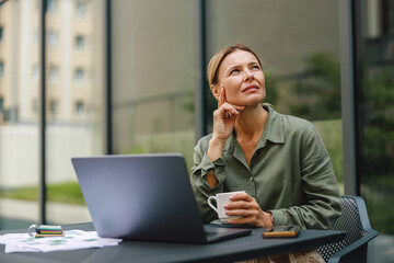 Wall Mural - Focused woman sales manager working on laptop and drinking coffee in terrace and looking at side