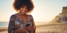 A Beautiful Black Girl Looking At Her Phone And Checking Social Media. Sunny Golden Light Beach And Ocean In The Background.