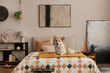 Cozy composition of bedroom interior with mock up poster frame, bed, orange bedding, corgi dog, wooden bedside table, black rack, stylish lamp, sculpture and personal accessories. Home decor. Template