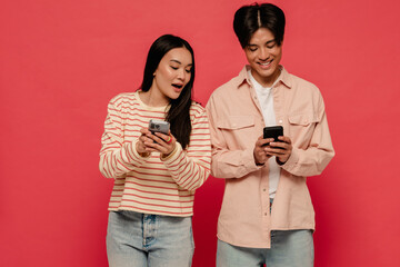 Asian woman looking at her boyfriend's mobile phone screen while standing isolated over red background