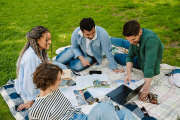 Wall Mural - Group of smiling students with laptop doing homework together while sitting in park