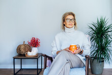 Middle Aged Woman Relaxing With Pumpkin Shaped Cup Of Hot Drink In Scandy Style Hygge Interior Home With Fall Mood Decor. Lady Dreaming, Enjoy Calm Mood Without Stress, Well Being Alone. Cozy Autumn