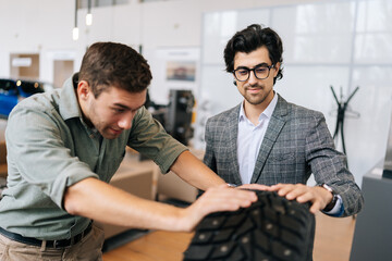 Wall Mural - Portrait of salesman in business suit talking about characteristic of tires product to cheerful customer male came to look at assortment represented in car dealership. Concept of auto maintenance.