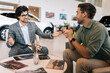 © dikushin - Happy male customer talking about terms of buying car with manager wearing business suit sitting at desk in auto dealership. Concept of choosing, buying new auto at showroom.