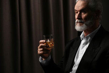 Poster - Senior man in suit holding glass of whiskey with ice cubes on brown background. Space for text