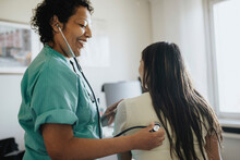 Side View Of Smiling Mature Female Doctor Examining Young Woman With Stethoscope In Clinic
