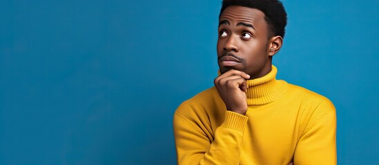 Canvas Print - Young African American man in blue sweater looking doubtful touching his face isolated on yellow background for advertisement