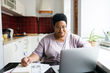 african plus size aged woman sitting in front of laptop planning purchases, making list before going