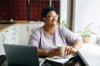 © shurkin_son - Cheerful happy smiling overweight mature black woman in spectacles sitting at kitchen table with white coffee cup in hands looking through window, enjoying hot drink, after working online on laptop