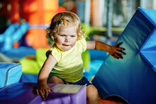 Happy Blond Little Toddler Girl Having Fun And Sliding On Indoor Playground At Daycare Or Nursery. Positive Funny Baby Child Smiling. Healthy Girl Climbing On Slide.